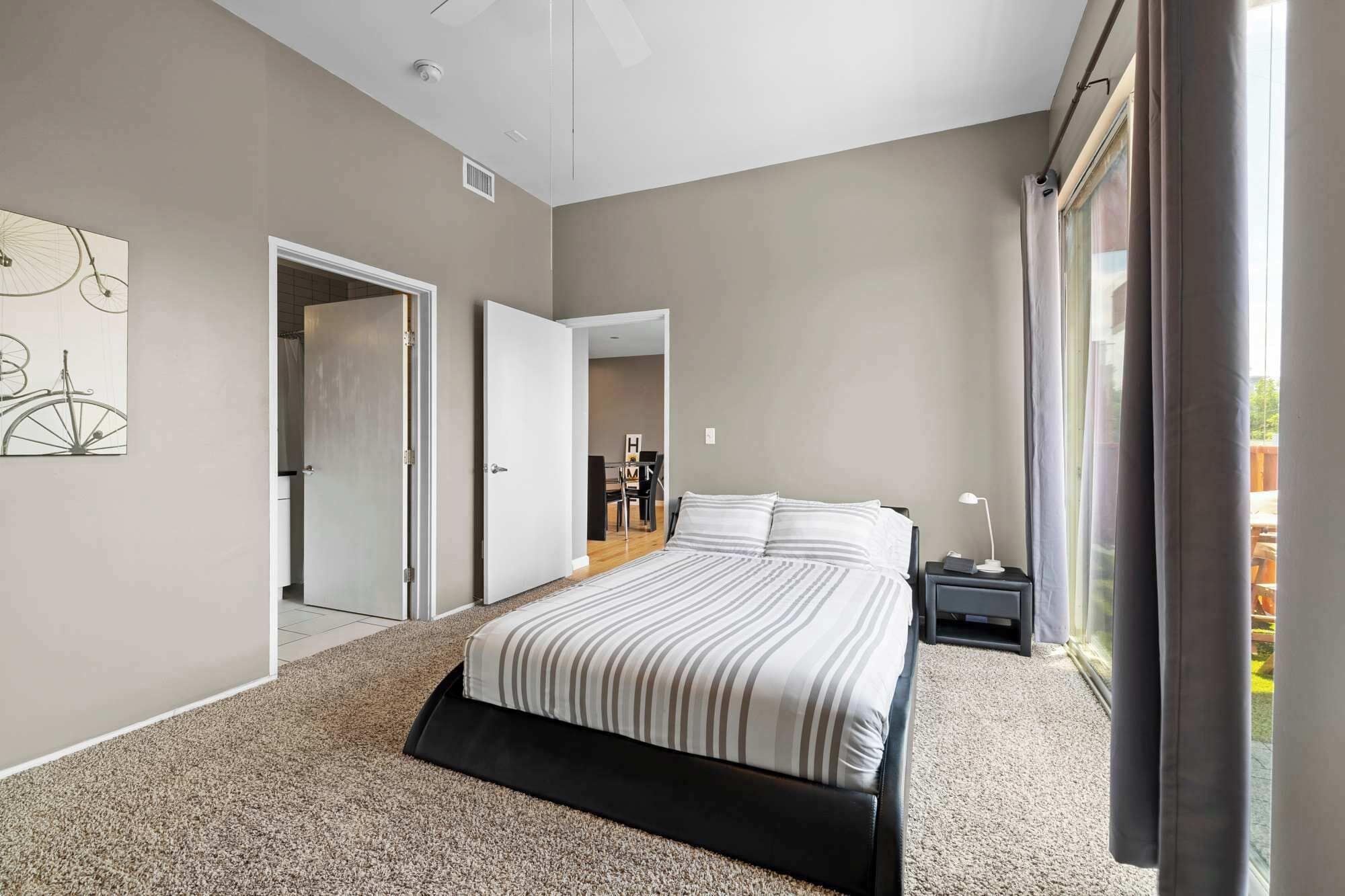 A modern bedroom with a large bed, a desk, and a chair. There is a door leading to another room and a window with curtains.