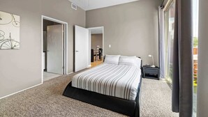 A modern bedroom with a large bed, a desk, and a chair. There is a door leading to another room and a window with curtains.