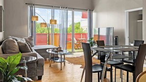 A modern living room with a sofa, a glass dining table, and chairs. There is a large window with curtains, a view of the outside, and a flat-screen TV on a stand.