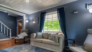 Un salon avec un canapé beige, un tapis à motifs, une fenêtre avec des rideaux bleus et un escalier en arrière-plan.