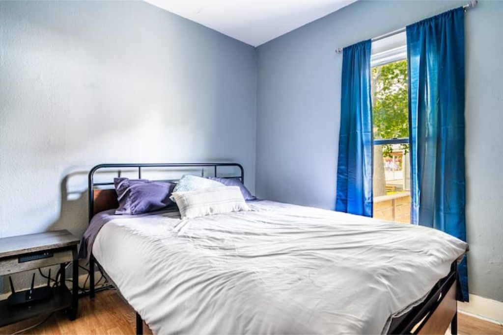 Una cama individual con colchón blanco y estructura metálica, un banco con cojín y una ventana con cortinas azules.