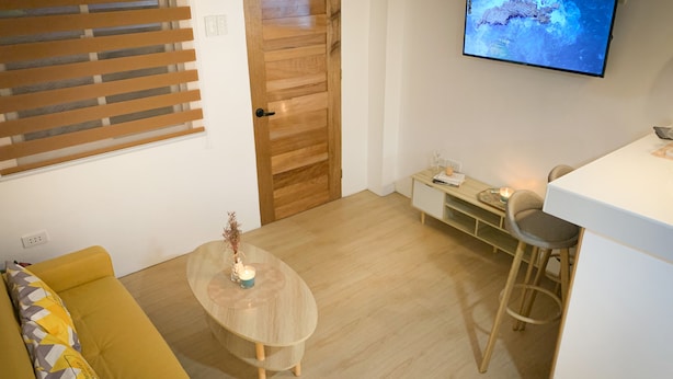 Ein modernes Wohnzimmer mit einem gelben Sofa, einem Holztisch, einem an der Wand montierten Fernseher und einer kleinen Schreibtischzeile.