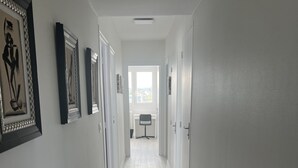 Un pasillo con paredes blancas, piso de madera clara y obras enmarcadas a la izquierda.