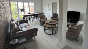 Una sala moderna con un sofá de cuero, una mesa redonda y un televisor. Se observa un comedor con una mesa y sillas al fondo.