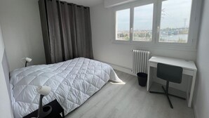 Un dormitorio con cama, escritorio, silla, radiador y ventana con vista a un puente y edificios.