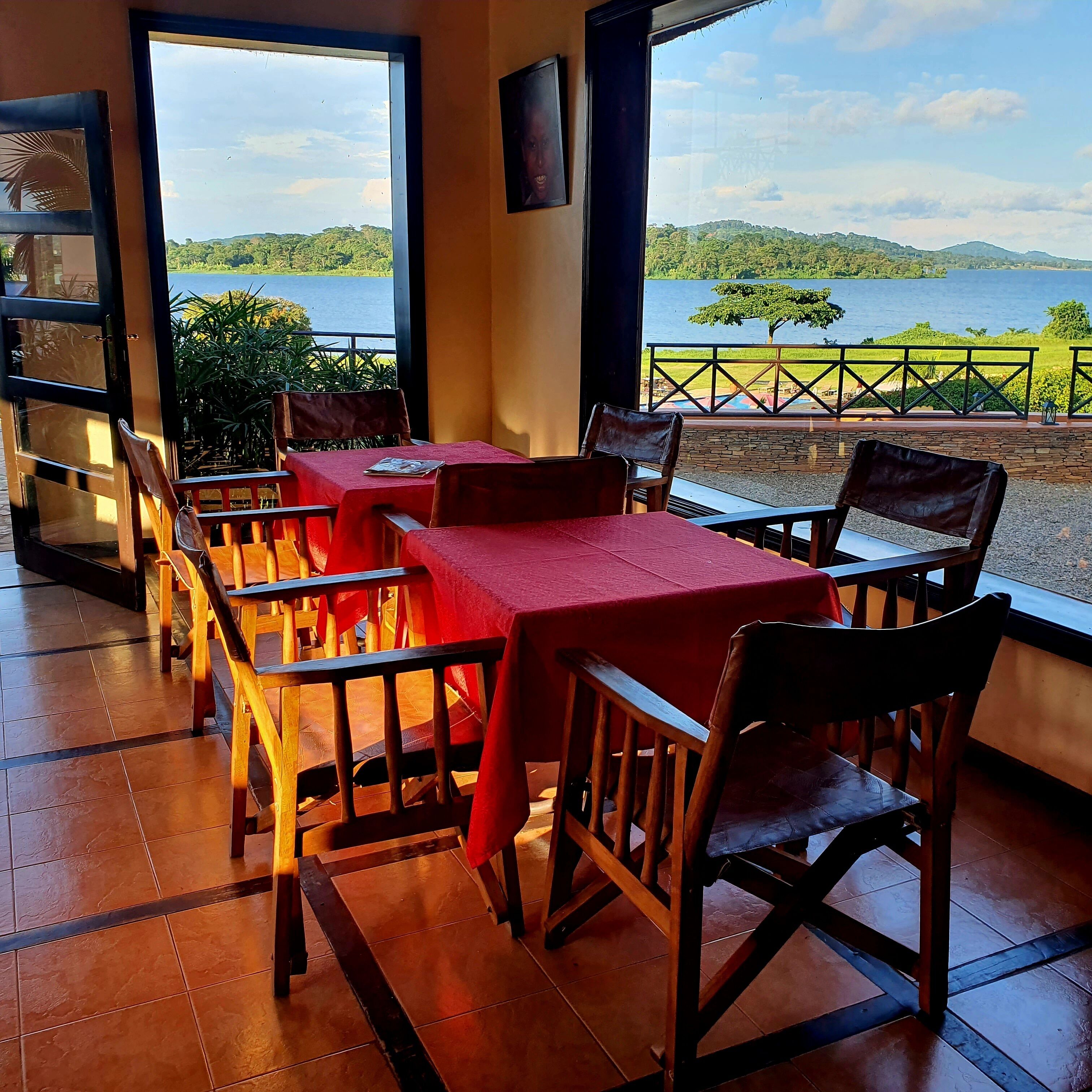 Um restaurante com mesas e cadeiras de madeira, toalhas de mesa vermelhas e vista para um lago e colinas.