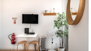 Et moderne værelse med et fladskærms-tv, et lille skrivebord, to træstole, en potteplante, en gulvventilator, en brandslukker og en træhylde med dekorative genstande.