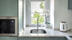 A kitchen with a stainless steel sink, a microwave, a white kettle, and a window with a view of greenery. - Studio in Central Lisbon by Lisbeyond (Lisboa)