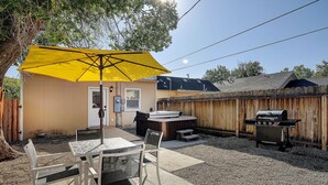 Ein Garten mit einer Terrasseneinrichtung, Stühlen, einem gelben Sonnenschirm, einem Whirlpool und einem Grill.