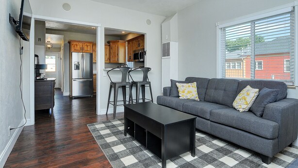 Ein modernes Wohnzimmer mit grauem Sofa, einem schwarzen Couchtisch und einem karierten Teppich. Durch einen offenen Durchgang ist eine Küchenzeile mit Holzschränken und Edelstahlgeräten erkennbar.