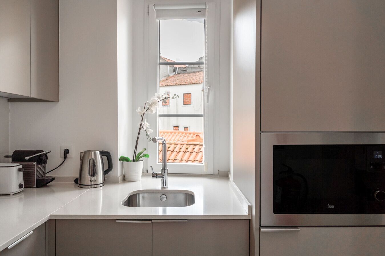 Una cocina moderna con un lavabo de acero inoxidable, una tetera, un tostador y vistas a un tejado de tejas rojas a través de una ventana.