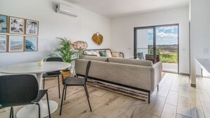 A modern living room with a sofa, a dining table with chairs, a large window, and a view of the outdoors. - Sun, Sand & Surf Apt in Aljezur by Lisbeyond (Aljezur)