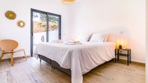 A bedroom with a bed, a chair, a bedside table with a lamp, and a view of the outdoors through a door. - Sun, Sand & Surf Apt in Aljezur by Lisbeyond (Aljezur)