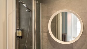 Une douche équipée d’un pommeau de douche à main et d’un flexible de douche, d’un miroir rond et d’une bouteille de shampoing.