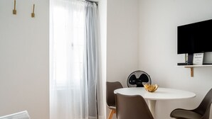 Une pièce moderne avec une table à manger ronde, deux chaises, un petit ventilateur, un téléviseur fixé au mur et une fenêtre ornée de rideaux légers.