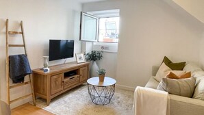 A compact living space with a wooden TV stand, a flat-screen TV, a small round table, a sofa, and a window with a view. - Stay in the heart of Alfama! | Cozy Apartment (Lisboa)