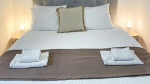 A neatly made bed with a grey upholstered headboard, white bedding, and two folded towels on a brown bed runner. - Stay in the heart of Alfama! | Cozy Apartment (Lisboa)