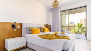 A modern hotel room with a large bed, a bedside table, a wall-mounted lamp, and a view of palm trees through a sliding glass door. - Oasis Mar Beach Apartment by Lisbeyond (Portimão)