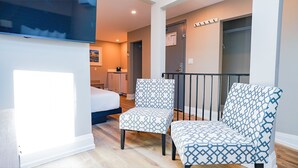 A modern hotel room with a bed, two patterned armchairs, a small table, and a balcony with a railing.