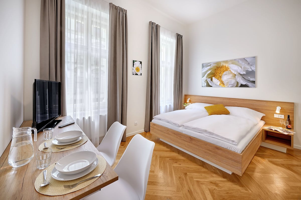 A hotel room with a wooden bed, a desk with a TV, and a dining area with a pitcher of water.