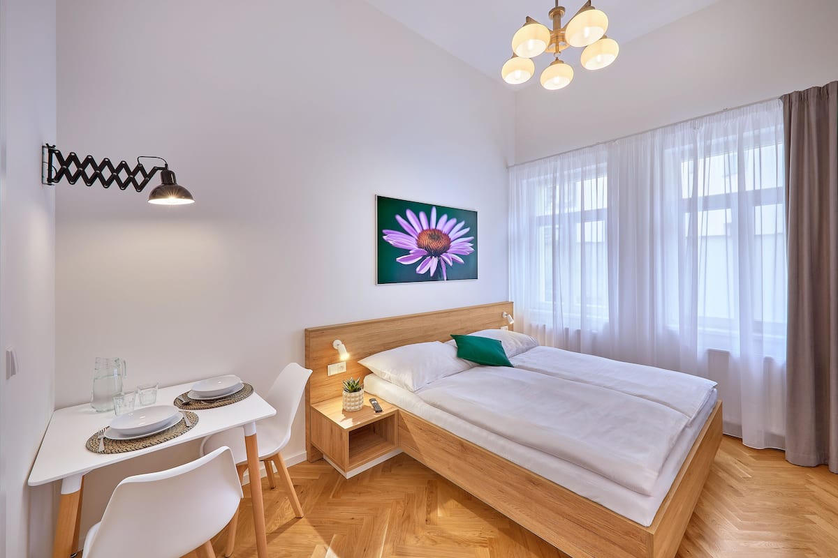 A modern bedroom with a wooden bed, white bedding, a desk with a lamp, and a painting of a flower on the wall.