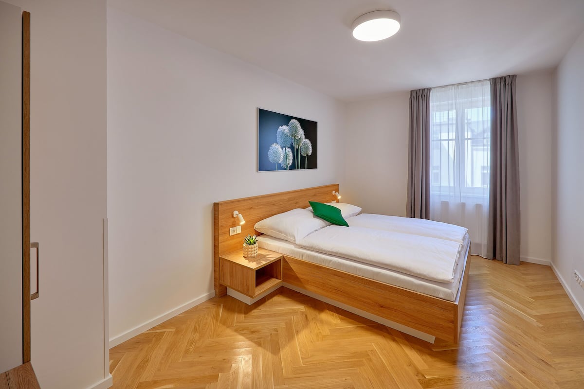 A modern bedroom with a wooden bed, a nightstand, a wall-mounted light, and a window with curtains.