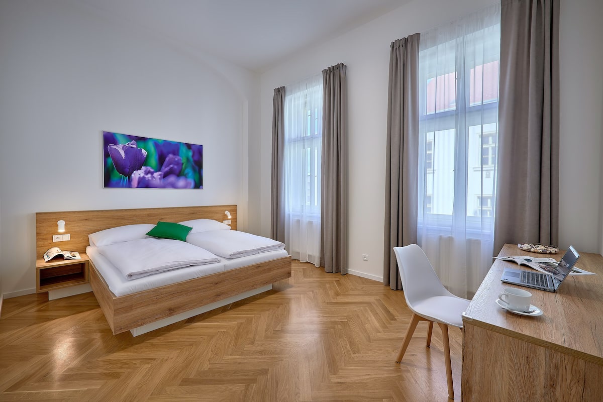 A modern hotel room with a wooden bed, a desk with a laptop, and a view of the city through large windows.