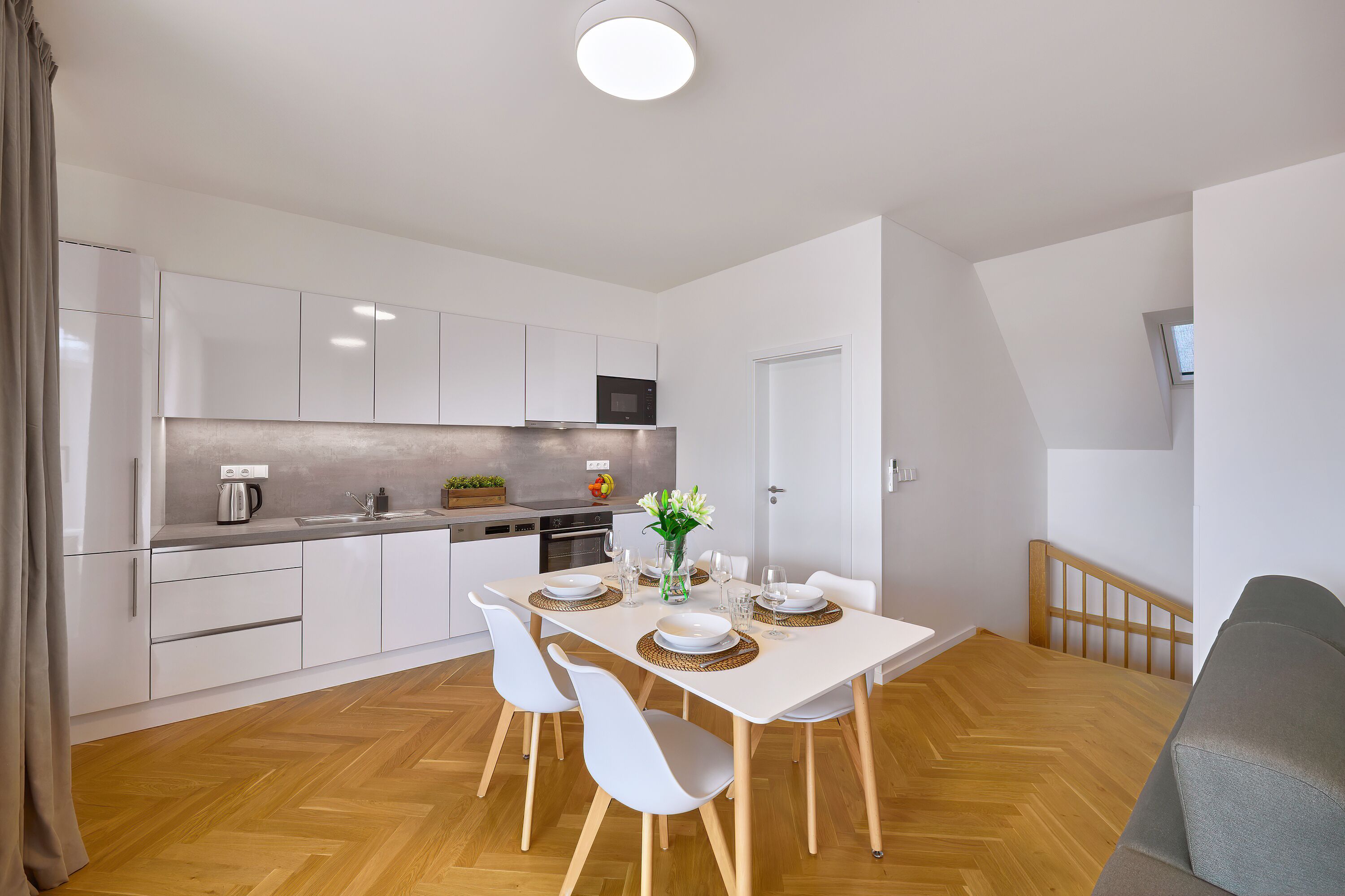A modern kitchen with white cabinets, a wooden herringbone floor, and a dining area with a white table and chairs.