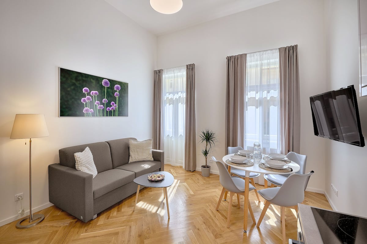 A modern living room with a grey sofa, a small round table, a dining table with chairs, a TV mounted on the wall, and a large window with curtains.