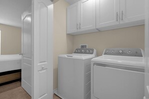 A laundry room with a washing machine and dryer, a door, and a bed in the background. - 4730 Lakeview Townhome with Pool Access Near Disney (Kissimmee)