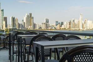 A rooftop terrace with tables and chairs, overlooking a city skyline.