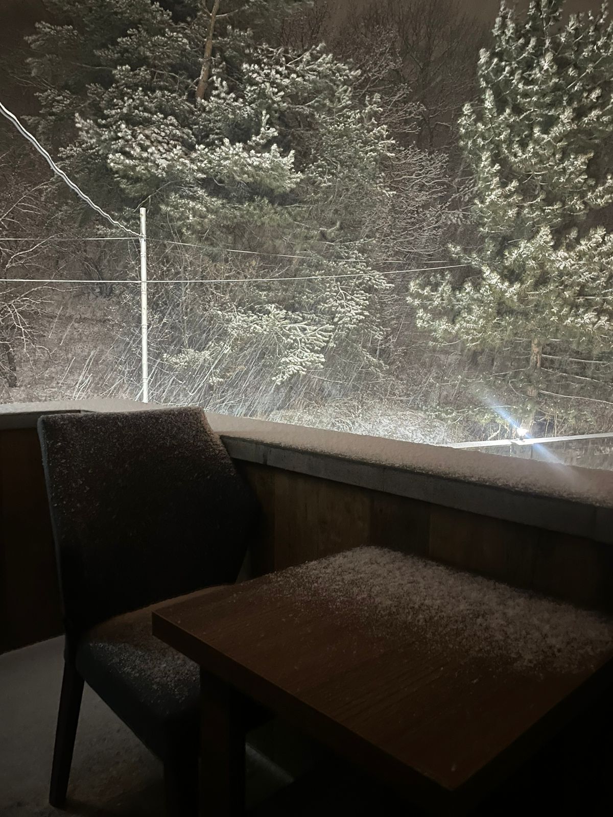 Una habitación tenuemente iluminada con una mesa de madera y una silla. Una ventana muestra un paisaje nevado con árboles y una línea de electricidad.