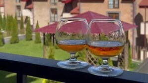 Two glasses of amber liquid on a balcony railing with a view of a building with red roofs and a clear blue sky. - Marrone Hotel Tsaghkadzor (Tsaghkadzor)