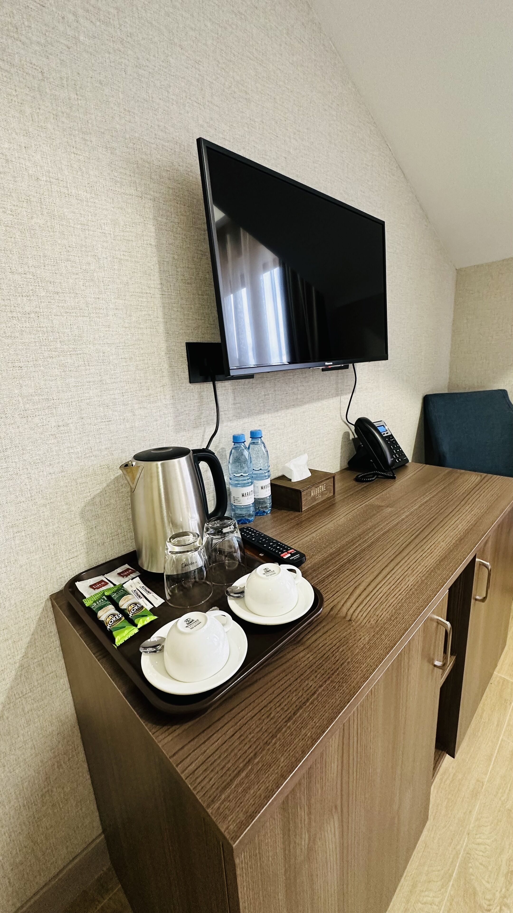 A hotel room with a wooden desk, a flat-screen TV mounted on the wall, a telephone, and a tray with coffee, water bottles, and tea packets.