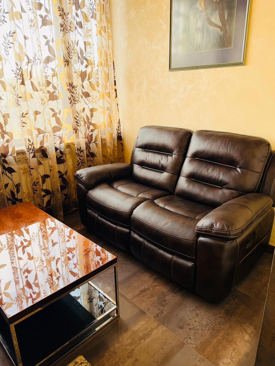 A brown leather sofa, a glass-top coffee table, and patterned curtains in a living room.