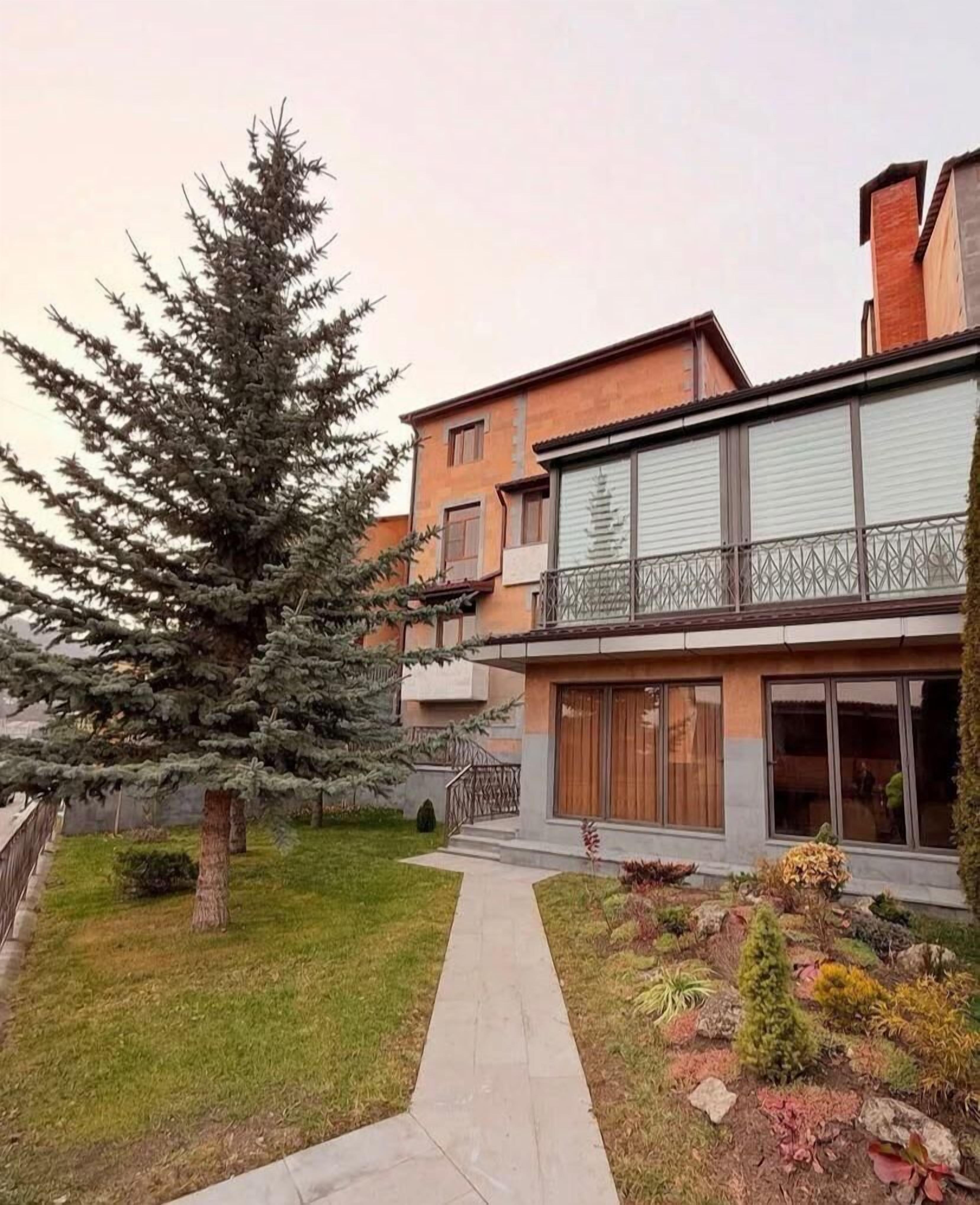A modern two-story residential building with a balcony, a large coniferous tree, and a well-maintained garden.