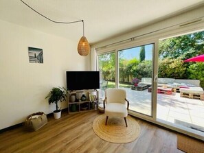 Una sala de estar con televisión, una silla, una planta y un amplio ventanal corredera que da a un patio exterior.