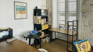 A cozy room with a wooden table, a bookshelf, a framed picture, and a window with shutters.
