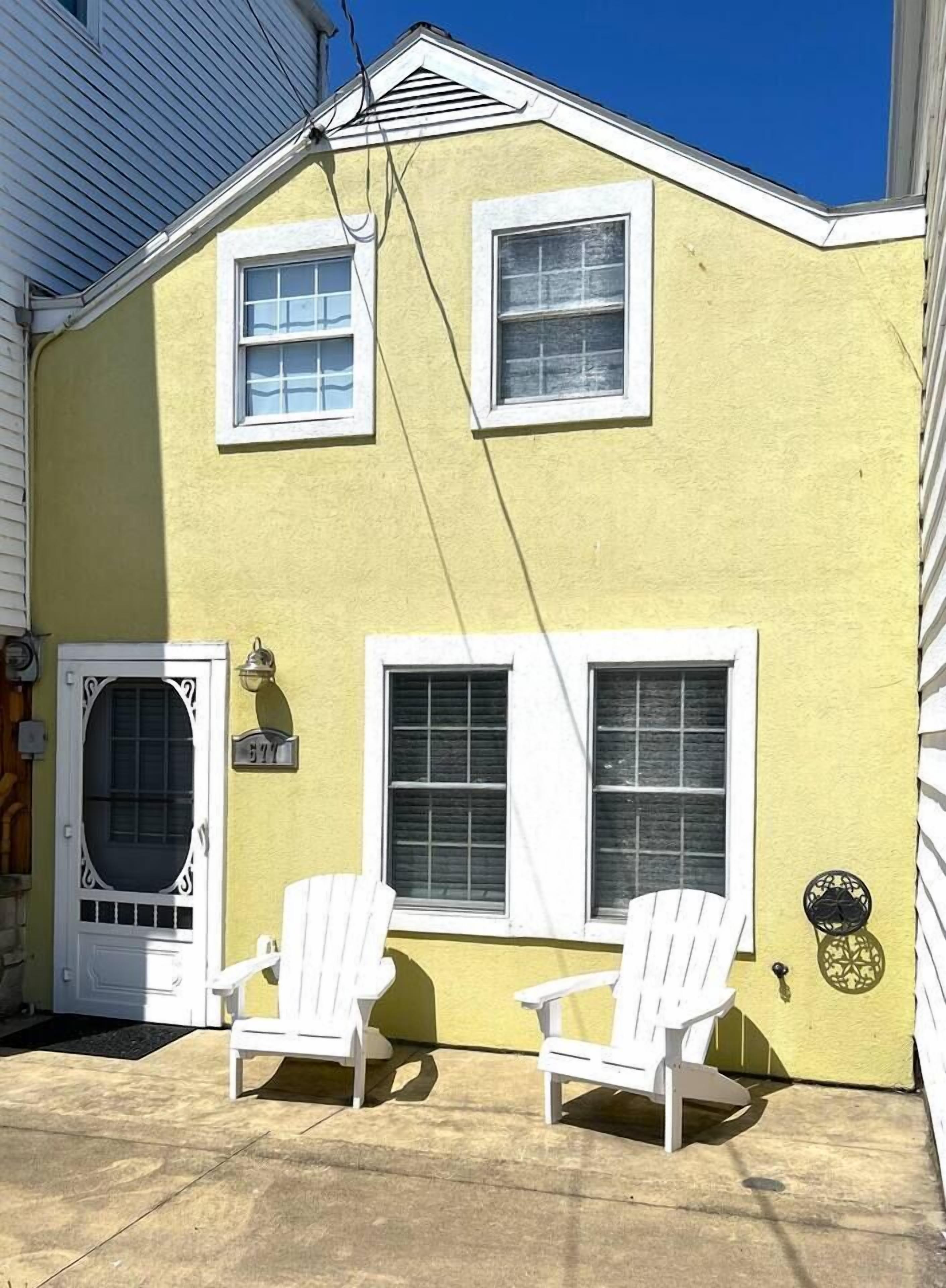 A yellow house with two white windows and a door, two white Adirondack chairs outside, and a small sign on the door.