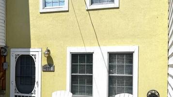 A yellow house with two white windows and a door, two white Adirondack chairs outside, and a small sign on the door.