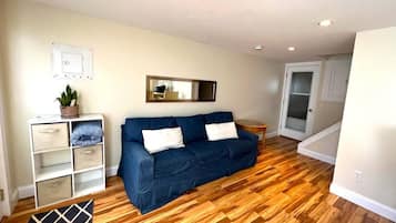 A living room with a blue sofa, wooden flooring, a small side table, and a white wall-mounted shelf.