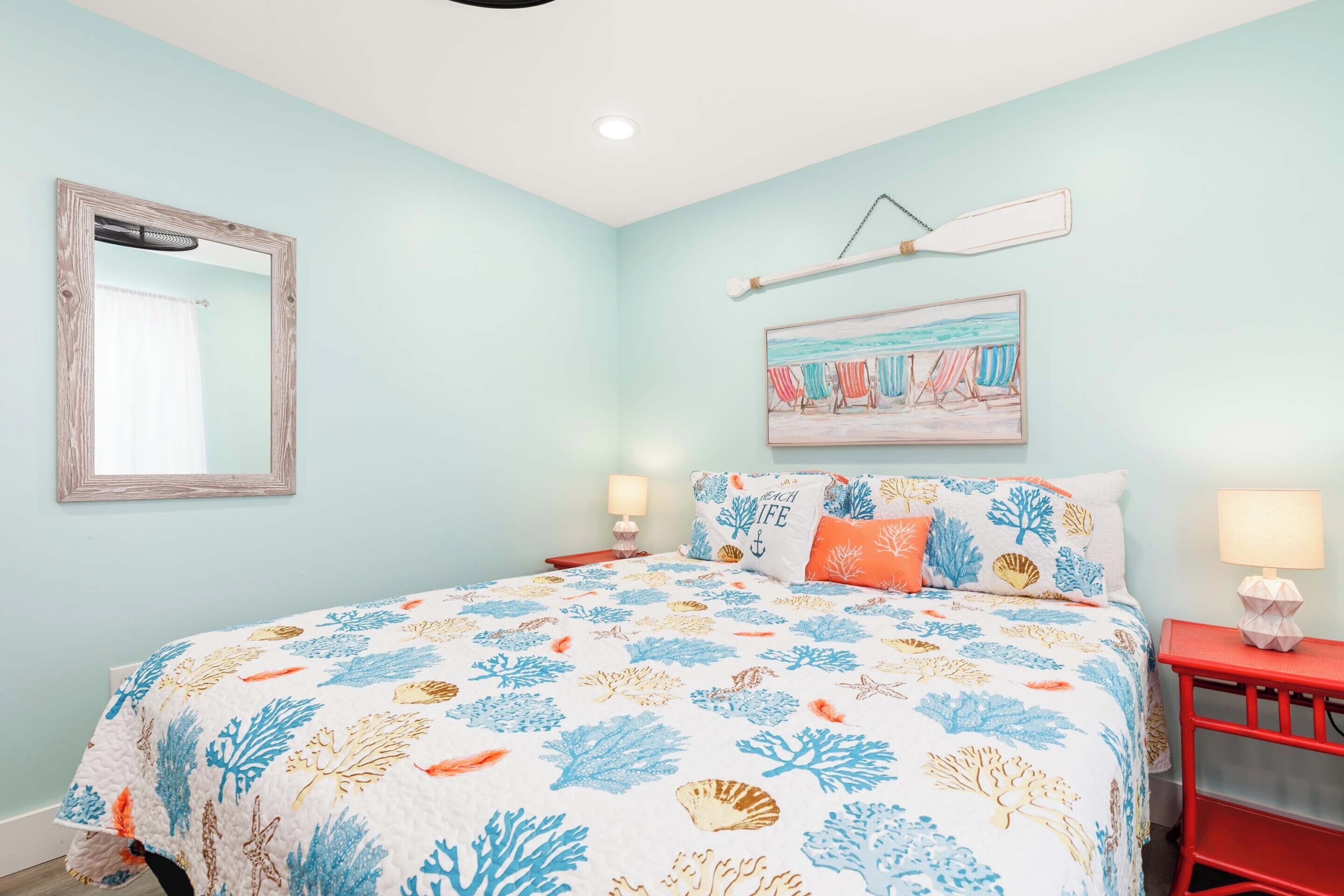 Un dormitorio con cama, dos mesitas de noche, un espejo en la pared, un cuadro con motivos playeros y un ventilador de techo.