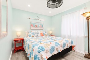Un dormitorio con cama, dos mesitas de noche, un ventilador de pared, una ventana con cortinas y una obra de arte con temática playera.