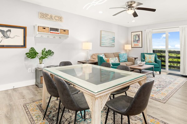 Uma sala de estar moderna com uma mesa de jantar de vidro, cadeiras escuras, um sofá bege e poltronas verde-água, além de uma grande janela com vista.