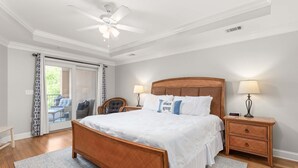 Un dormitorio con una cama de madera, dos mesitas de noche con lámparas, un ventilador de techo y vistas a una zona de estar exterior a través de un ventanal.