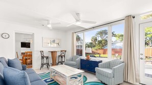 A spacious living room with a large sofa, a coffee table, and two armchairs. There is a dining area with a table and chairs, and a view of the backyard through large windows. - Tybrisa Treasure by Tybee Vacation Rentals (Tybee Island)