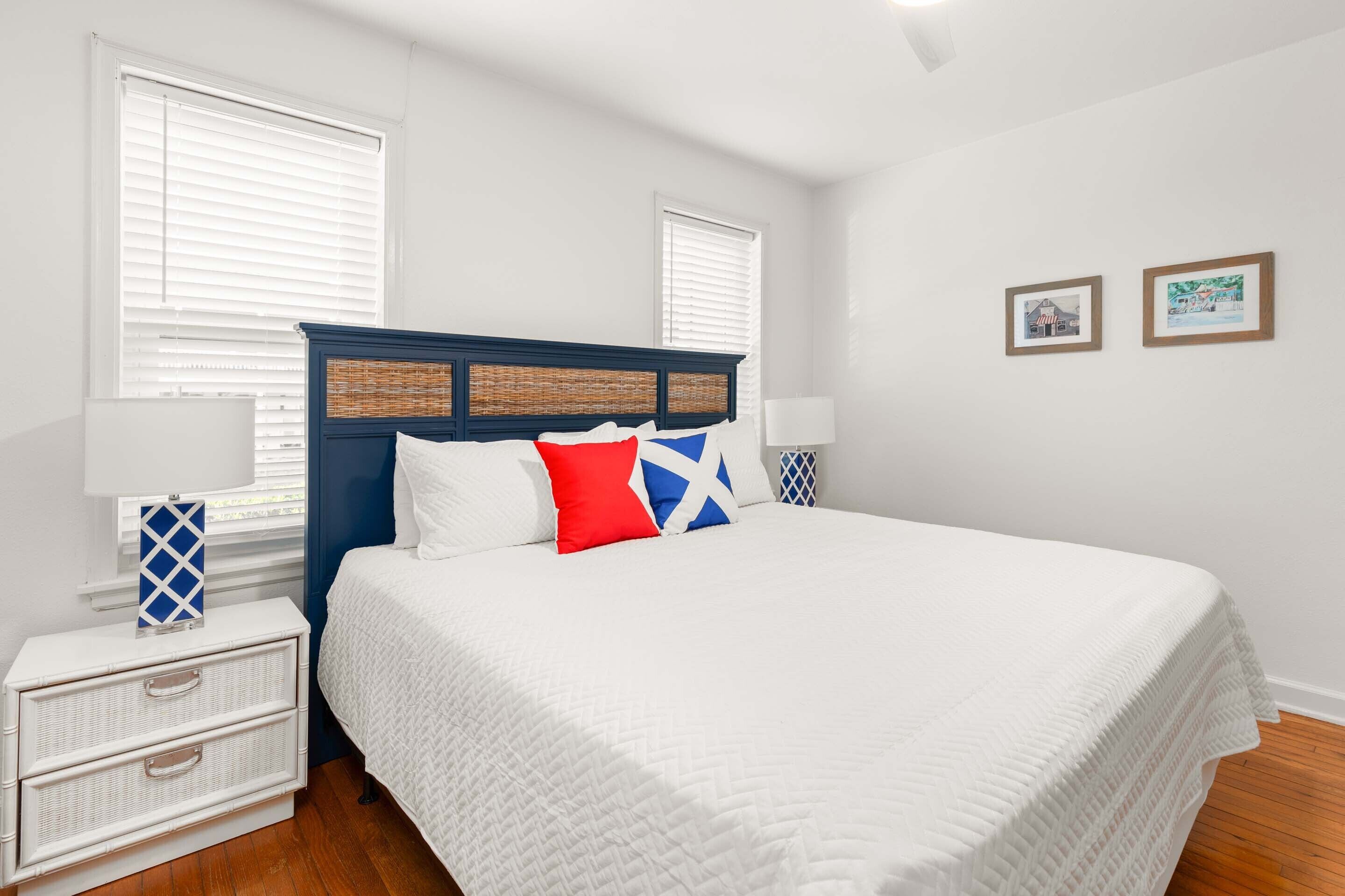 A bedroom with a large bed, white bedding, a blue headboard, and two windows with blinds.
