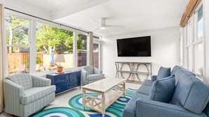 A spacious living room with a large window, a ceiling fan, a flat-screen TV, and a blue sofa. - Tybrisa Treasure by Tybee Vacation Rentals (Tybee Island)