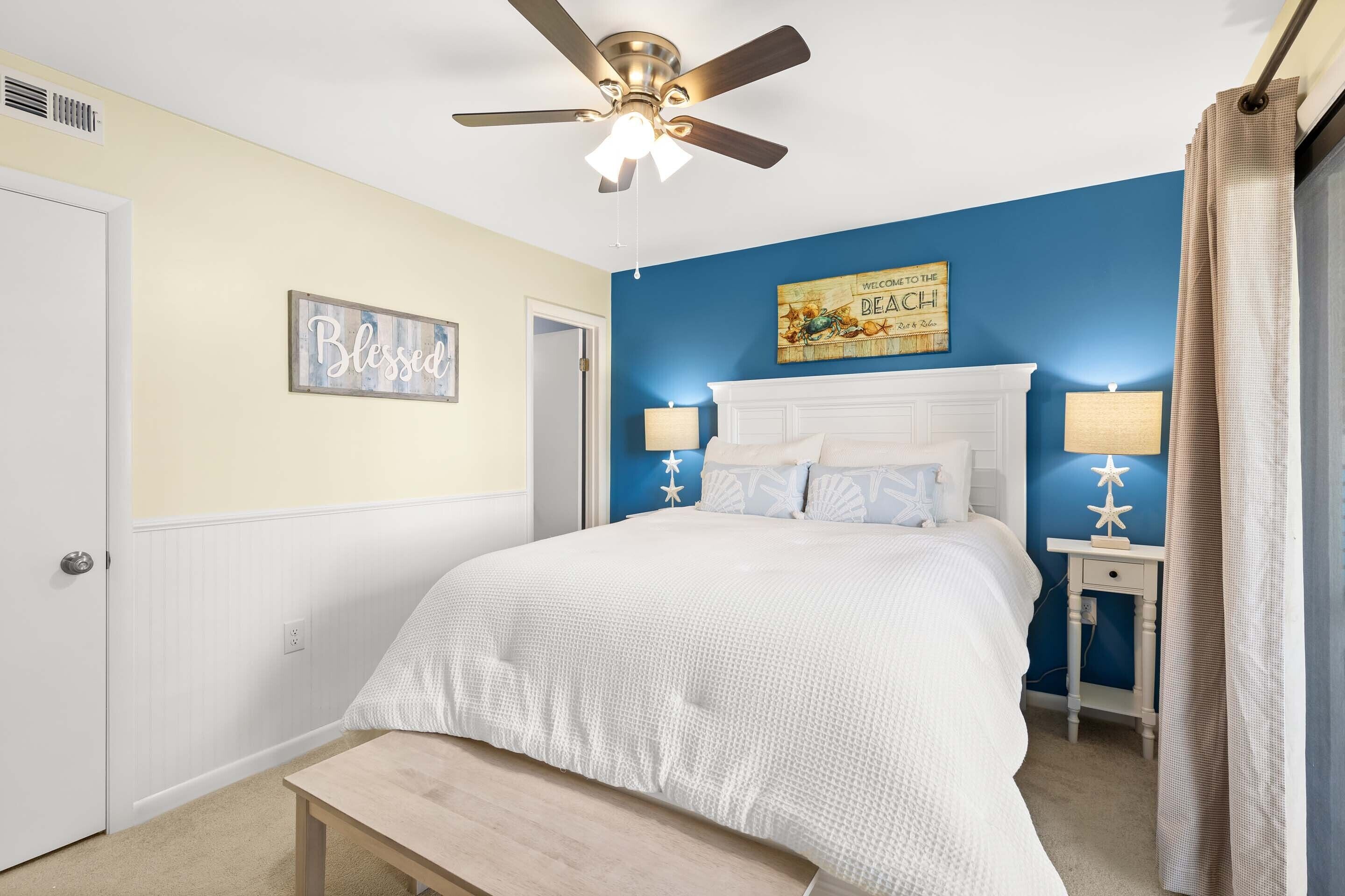 A bedroom with a bed, a ceiling fan, a bedside table with a lamp, and a wall with a 'Welcome to the Beach' sign.