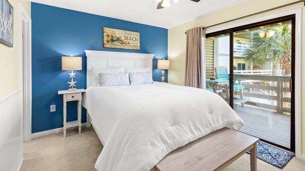 A bedroom with a large bed, a ceiling fan, a nightstand, a lamp, and a view of the outdoor deck. - Lighthouse Point 33A by Tybee Vacation Rentals (Tybee Island)